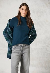 Tealfärgad ribbad tröja med hög krage, tillsammans med grå jeans. En grön och blå rutiga flanellskjorta ligger över axlarna.