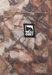 Földszínekkel ellátott, kamuflázs mintázatú anyag, mellzsebbel. Fekete címke fehér medve grafikával és "OBEY" felirattal.