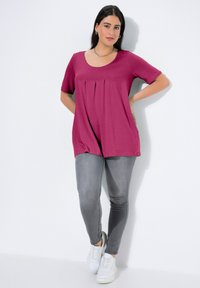 Magenta krótka bluzka z krótkim rękawem, z plisami z przodu, połączona z szarymi dopasowanymi dżinsami i białymi trampkami. Gładka tkanina o luźnym kroju.