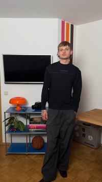 Młody mężczyzna w czarnej bluzie i szarych spodniach stoi w salonie z telewizorem zamontowanym na ścianie, pomarańczową lampą oraz niebieską półką z książkami i piłką do koszykówki.