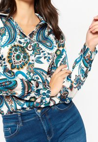 Kvinna som bär en färgglad skjorta med paisleymönster, nedstoppad i blå denimjeans, poserar med ena armen korsad och den andra upplyft.