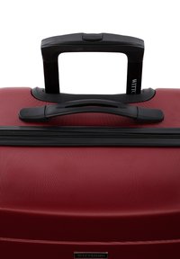 Maleta dura roja con una superficie texturizada, mango retráctil negro y cierre de cremallera. Contiene un logotipo de la marca en la parte frontal.