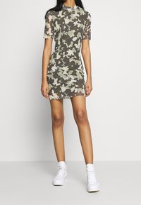 Kamouflage-mönstrad miniklänning i genomskinligt tyg med korta ärmar och hög halsringning; kombinerad med vita sneakers mot en neutral bakgrund.