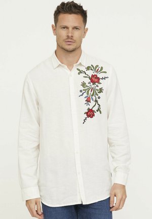 Chemise en lin blanc à manches longues, avec des broderies florales vives rouges et vertes sur le côté gauche et un devant boutonné.