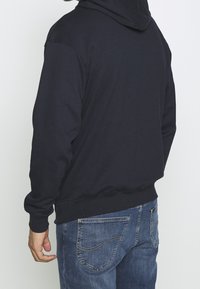 Černá mikina s kapucí vyrobená z měkké tkaniny, s dlouhými rukávy a žebrovaným lemem, nošená s modrými džíny.