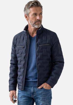 En midaldrende mand med gråt hår og skæg bærer en marineblå quiltet jakke over en blå sweater og jeans, og holder en smartphone i den ene hånd.