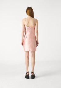 Robe nuisette en satin rose avec des fines bretelles, présentant une silhouette ajustée et une couture à l'arrière. Associée à des sandales à talons épais noires.