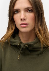 Hoodie verde oliva con cordini con punte dorate, tessuto morbido, colletto a coste e una vestibilità comoda attorno al collo.