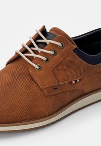 Brune suedesko med perforert tekstur, marineblå detaljer, rund tå og beige såle. Har kontrastfargede snøring og lyse lisser.