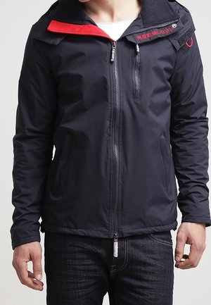 Homme portant une veste noire zippée avec un col intérieur rouge et une petite poche avant, associée à un jean foncé.