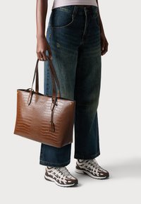 Persoon in wijde blauwe jeans en witte sneakers, die een grote bruine leren schoudertas met textuur bij de handvatten vasthoudt.