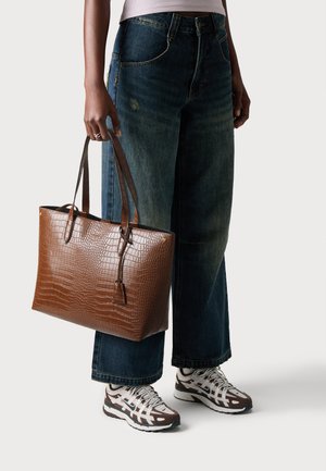 Person wearing wide-leg blue jeans and white sneakers, holding a large brown textured leather tote bag by the handles.