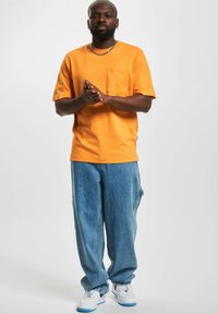 Oransje bomull t-skjorte med brystlomme, kombinert med løse blå denimbukser. Modellen har på seg hvite sneakers med blå detaljer.