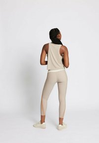Beiger Sporttanktop mit lockerer Passform, kombiniert mit enganliegenden Leggings, die seitliche Ausschnitte und eine weiche Textur aufweisen. Schuhwerk: hellfarbige Sneaker.