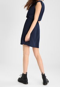 Marineblaues ärmelloses Kleid mit tailliertem Schnitt, fließendem Rock und glatter Textur, kombiniert mit schwarzen Schnürstiefeletten.