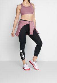Jasnofioletowy stanik sportowy, czarne legginsy z białym nadrukiem, różowe i białe sneakersy. Gładka tkanina, dopasowany krój i warstwowy wygląd.