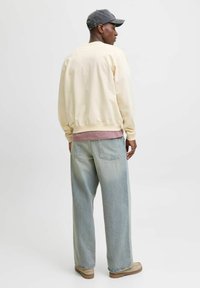 Creme sweatshirt med ribbede manchetter, lagd over en lyserød skjorte. Parret med lyseblå vide jeans og brune sko, sat mod en neutral baggrund.