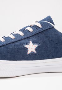 Modré plátěné tenisky s bílou gumovou podrážkou, na boku se starou nášivkou v bílé barvě a růžovými puntíky, a b ílými tkaničkami.