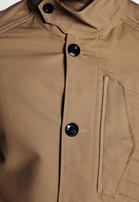 Gros plan sur une veste beige montrant trois boutons noirs, une poche poitrine rectangulaire verticale avec des détails cousus et un col avec fermeture par bouton.