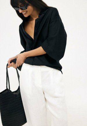 Woman wearing black oversized shirt and white pants holds a black woven tote bag, smiling and looking down.