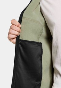 Svart zip-jacka med en mjuk grön fleecefoder och en intern svart ficka för förvaring. Texturerade tygdetaljer synliga.