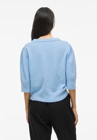 Suéter azul claro com um corte solto, mangas curtas e um decote largo. Textura suave e punhos canelados. Combinado com calças pretas.