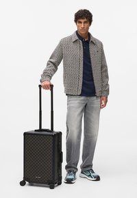 Fekete mintás gurulós bőrönd tyúklábmintás díszítéssel; tartós anyag, kihúzható fogantyú, négy kerék. Modell texturált dzsekiben és szürke farmernadrágban.