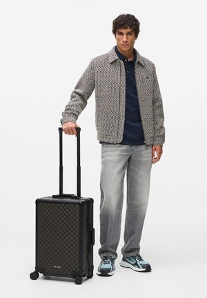 Fekete mintás gurulós bőrönd tyúklábmintás díszítéssel; tartós anyag, kihúzható fogantyú, négy kerék. Modell texturált dzsekiben és szürke farmernadrágban.