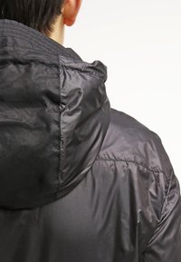 Veste matelassée noire dotée d'une capuche, d'une texture lisse et de coutures apparentes le long des coutures. Le dos présente un design épuré et minimaliste.