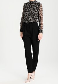 Blusa nera con motivo floreale, maniche lunghe in tessuto trasparente e collo alto, abbinata a pantaloni neri a vita alta e tacchi alti color nude.