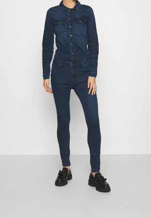 Kvinde iført mørkeblå tætsiddende denimjakke og jeans med sorte chunky loafers, stående mod en ensfarvet lys baggrund.