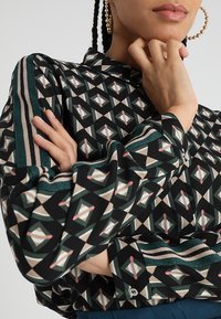 Blusa nera a fantasia con forme geometriche in verde, crema e rosa, con polsini a righe e chiusura a bottoni.