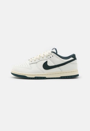 Hvid lav-tops Nike sneaker med mørkegrøn swoosh, mørkegrøn sål og lys beige mellemsål på en ensfarvet baggrund.