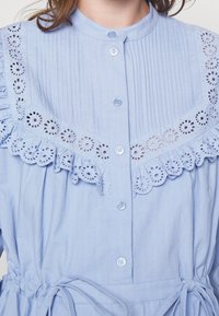 Ljusblå blus med texturerad tyg, plisserad design, knappstängning och en dekorativ öljettfrans längs kragen.