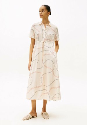 Vestito color crema con colletto abbottonato, maniche corte e vita stretta, decorato con un motivo a catena rosa e nero. Indossato con scarpe slip-on intrecciate.