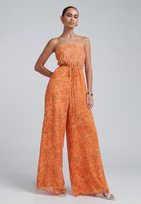 Trägersaler orangefarbener Jumpsuit mit floralem Muster, gefaltetem Oberteil und weitem Bein. Das Material wirkt leicht und strukturiert.