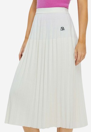 Frau trägt einen hoch taillierten, weißen, plissierten Midirock mit einem kleinen schwarzen, bestickten Logo auf der linken Seite und ein pinkfarbenes ärmelloses Oberteil.