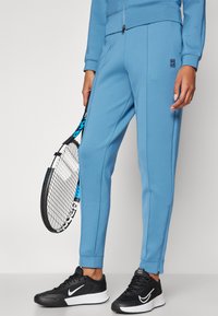 Blauwe sportbroeken van zachte stof, slim-fit ontwerp, met zijzakken en taps toelopende benen, gecombineerd met zwarte sportschoenen en een tennisracket.