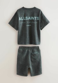 T-paita tummanvihreä pesty, jossa on "ALLSAINTS" ja japaninkielistä tekstiä, yhdistettynä yhteensopiviin joustovyötäröshortseihin, joissa on takatasku ja logoteksti.