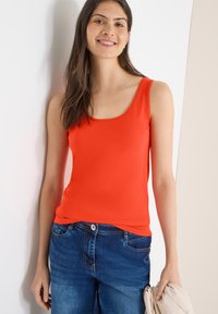 Femme souriante aux longs cheveux bruns portant un haut orange vif sans manches et un jean bleu, tenant une veste beige.