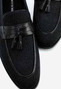 Zwarte loafers van een geweven materiaal met textuur, voorzien van een gladde zwarte leren band en kwastaccenten. Rond neuzen en een laag profiel ontwerp.