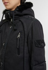 Svart parkas med hette foret med falsk pels, frontknapper og flere lommer. Har kontrastsøm og en glidelåslomme på brystet.