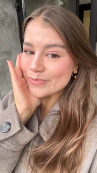 Lys brun ullkåpe med knappedetalj, langt bølgete brunt hår, subtil rosa sminke, fregner og gull hoops. 