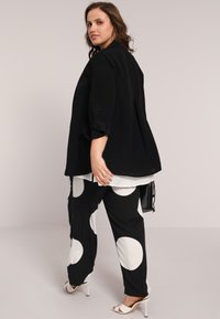 Veste noire ample à manches trois quarts, associée à un pantalon à pois noir et blanc ; le modèle porte des talons blancs.