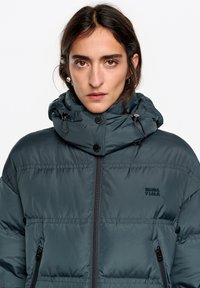 Kurtka puchowa z ciemnoszarego materiału z wysokim kołnierzem, zamkiem błyskawicznym z przodu i dwoma kieszeniami na zamek. Posiada haft z logo oraz regulowany kaptur.