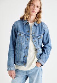 Hele sinine teksatagi, millel on ees nööbid, kaks rinnataskut ja pehme tekstuur. Kandes valge graafilise t-särgi peal. Allpool on nähtavad teksapüksid.