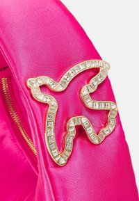 Różowa satynowa tkanina z broszką w złotym obrysie, ozdobioną przezroczystymi, prostokątnymi klejnotami, w kształcie stylizowanego zwierzęcia. Widoczny zamek.