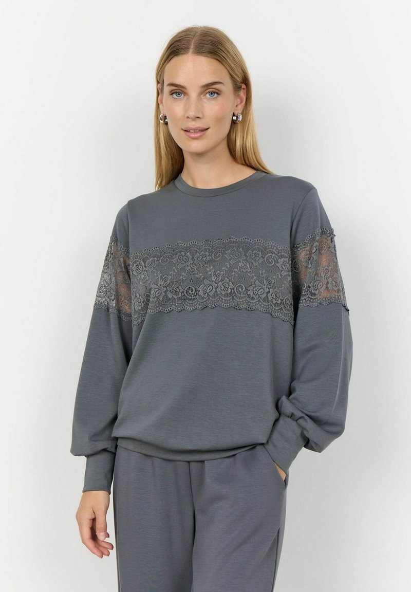 Sudadera gris de manga larga, con un borde de encaje en el pecho y las mangas superiores. Tela suave y ligera con un corte relajado.