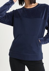 Mörkblå sweatshirt med en sammetslen överpanel och framficka. Långa ärmar, rundad halsringning och mjuk textur. Avslappnad passform.