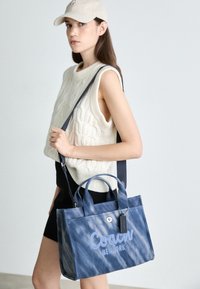 Blauwe leren tote bag met "Coach New York" borduursel, dubbele handvatten en een verstelbare schouderband; gladde textuur met een subtiele glans.